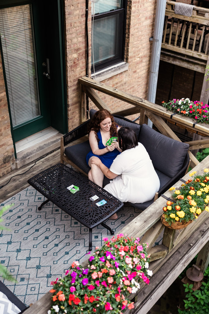 Two women sit closely on a balcony couch outside their home in Wicker Park, surrounded by flowers during their summer engagement session