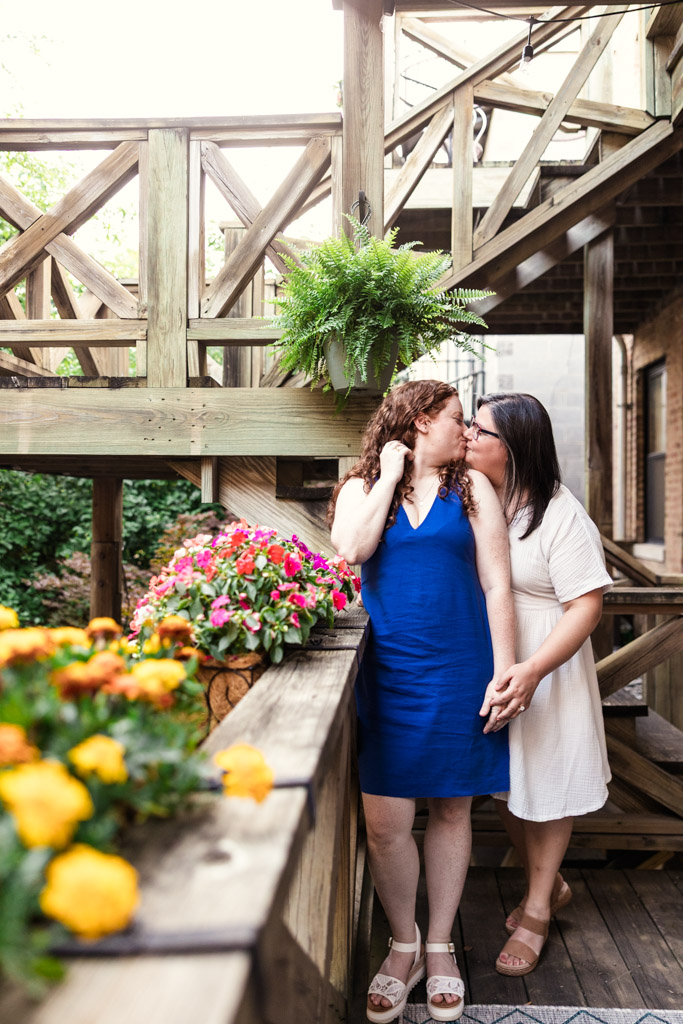 Two women hold hands and kiss on their wooden deck surrounded by colorful flowers and greenery during their Wicker Park engagement session