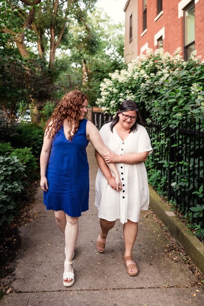 Two women smiling and holding hands while walking on a sidewalk near green plants and brick buildings during their Wicker Park engagement session