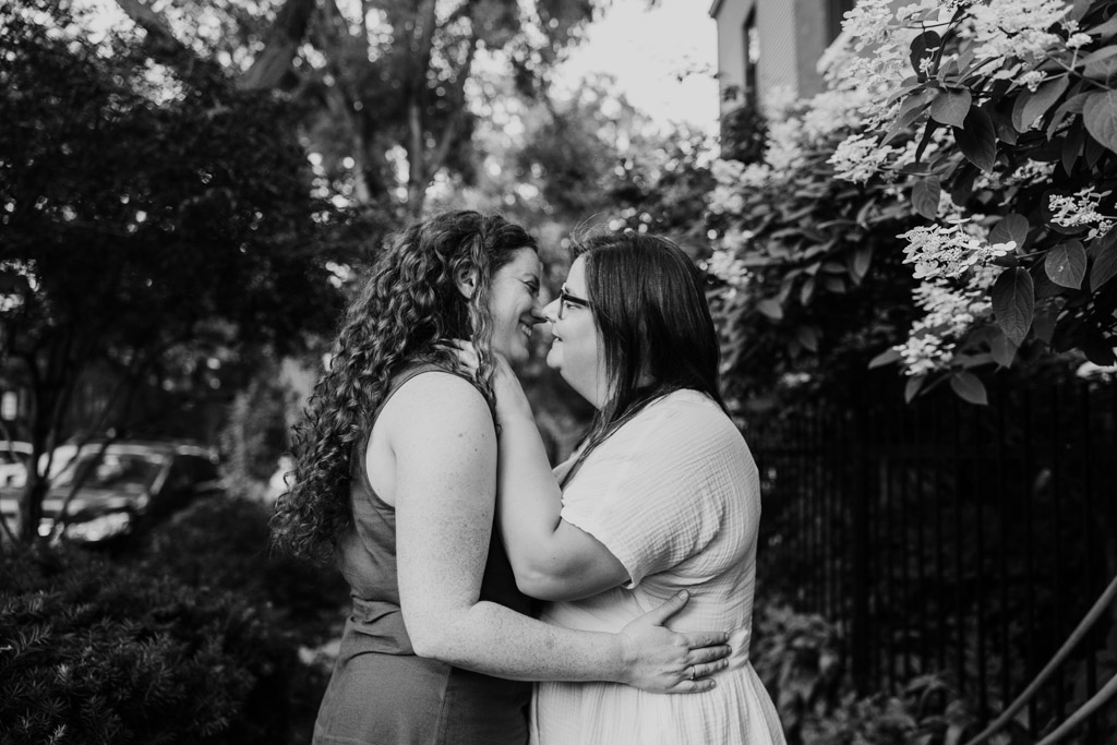 Black and white photo of two women smiling, standing close together, holding each other in a leafy, outdoor setting during their Wicker Park engagement session