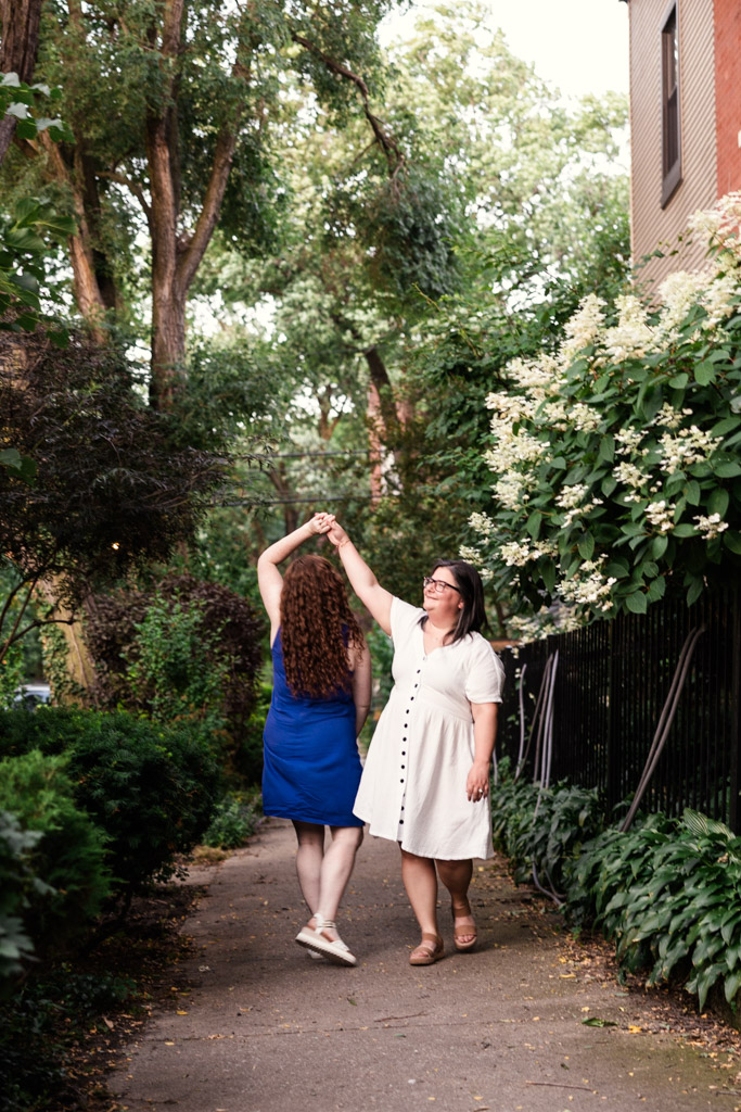 Engaged couple dances on a sidewalk surrounded by green plants and trees on a summer day, during their Wicker Park engagement session