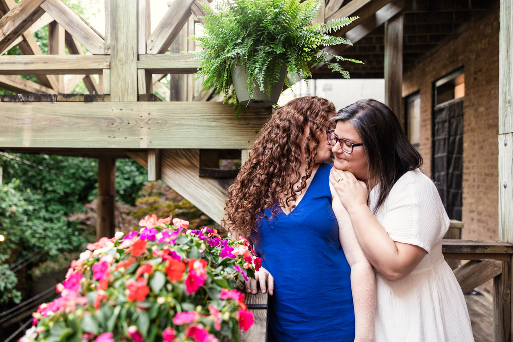 Woman kisses her partner's cheek on a wooden balcony filled with colorful flowers and green plants, during their Wicker Park engagement session