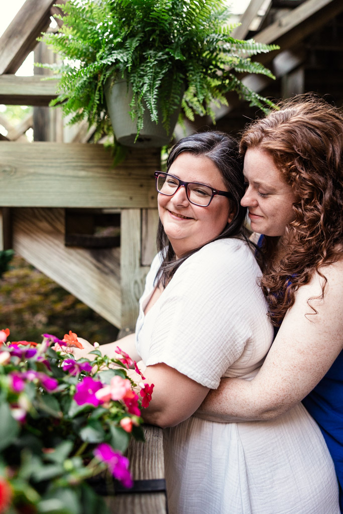 Two women embrace and smile by colorful flowers and wooden steps, with a fern hanging overhead, during their Wicker Park engagement session