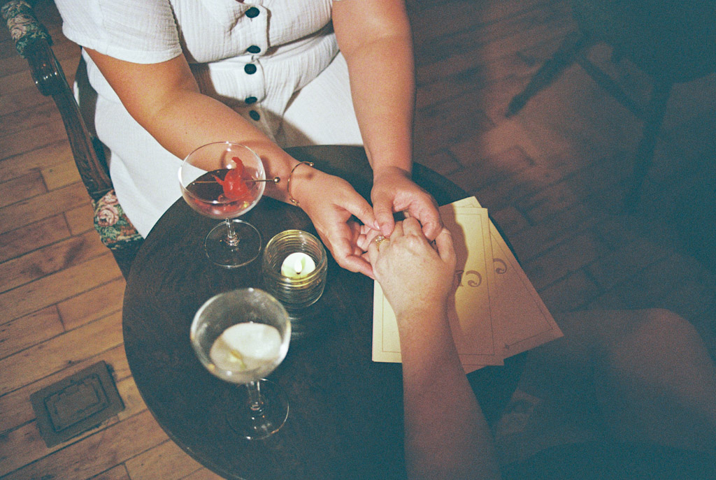 Two people hold hands at a small table with drinks, a candle, and menus, seen from above at Clara cocktail bar during summer engagement session