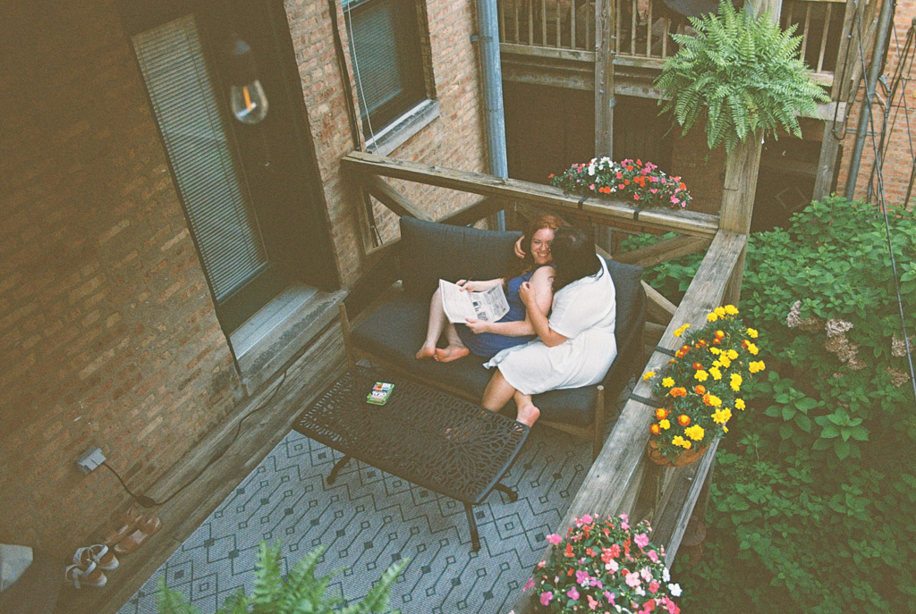 Two people relax on a balcony couch surrounded by flowers, reading and enjoying each other's company during their Wicker Park engagement session