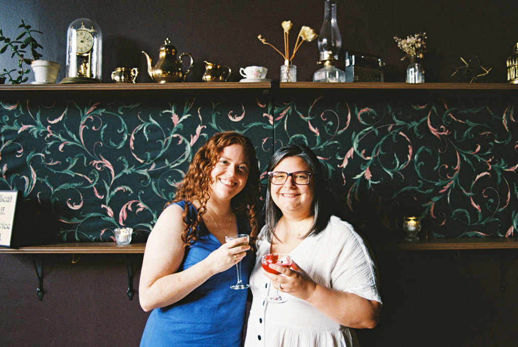 Film portrait of engaged couple smiling, holding drinks, standing in front of a floral-patterned wall with shelves and decorations inside Clara cocktail bar in Wicker Park