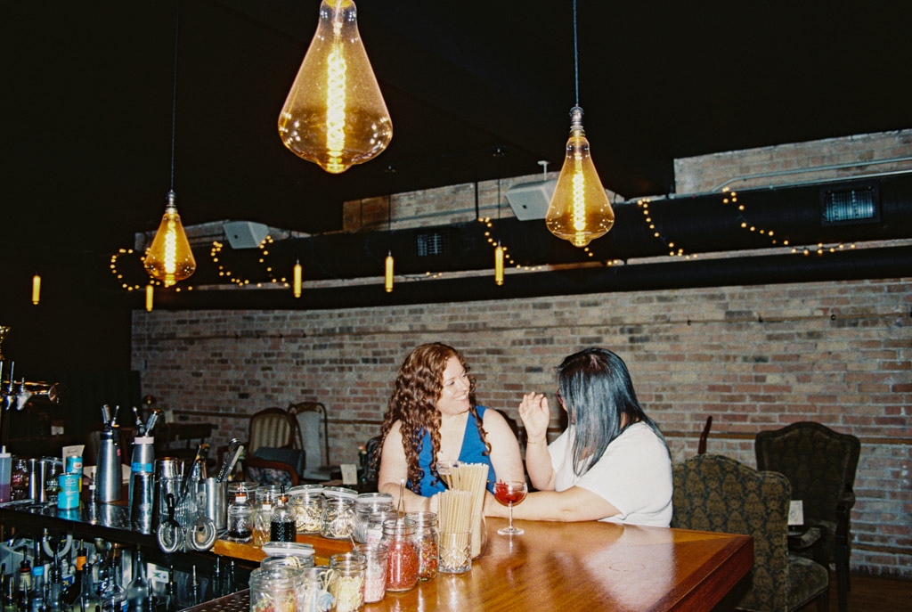 Film photo of wo women talking at the bar under warm hanging lights inside Clara in Wicker Park during summer engagement session
