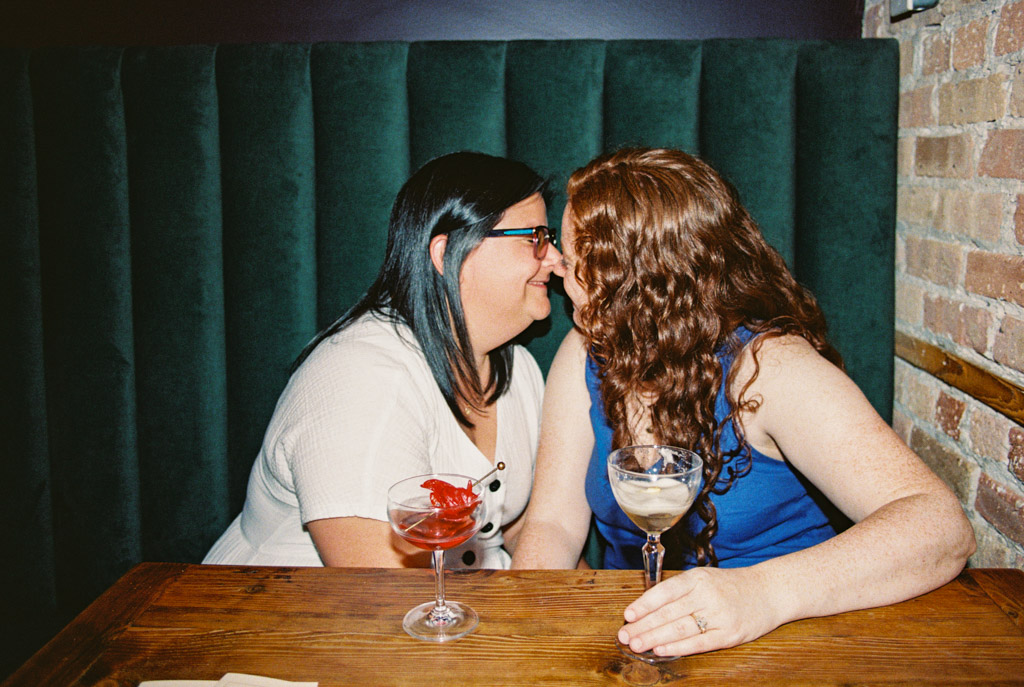 Two women sit close together at a table with drinks, smiling and about to kiss in a cozy booth during their Wicker Park engagement session