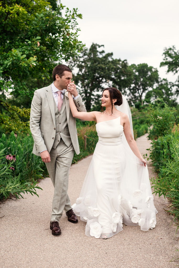 Groom kisses bride's hand as they walk down a Lincoln Park path after their intimate Alfred Caldwell Lily Pool elopement