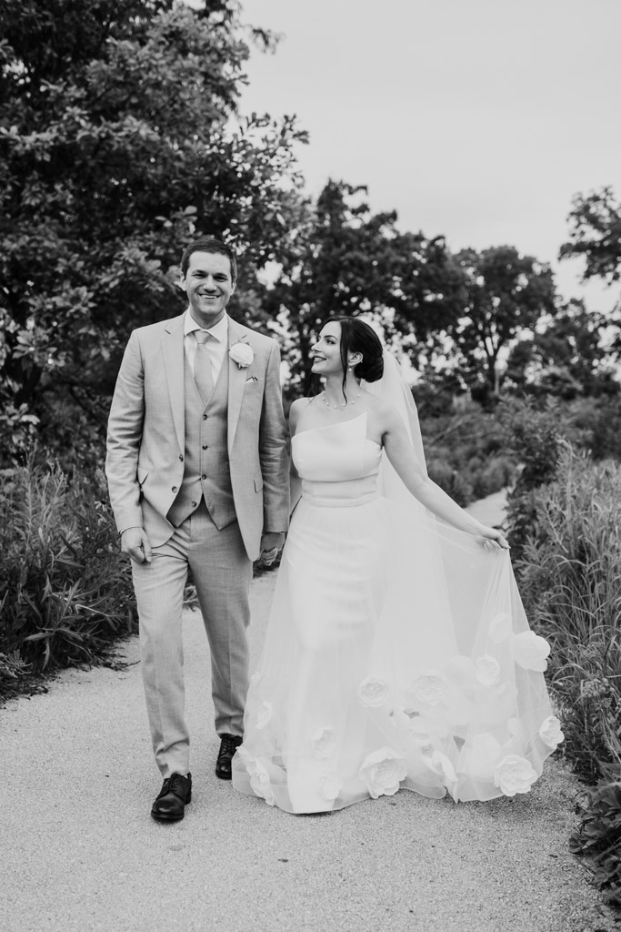 Black and white photo of bride and groom walking hand-in-hand on a Lincoln Park path outdoors, smiling after their intimate Alfred Caldwell Lily Pool elopement