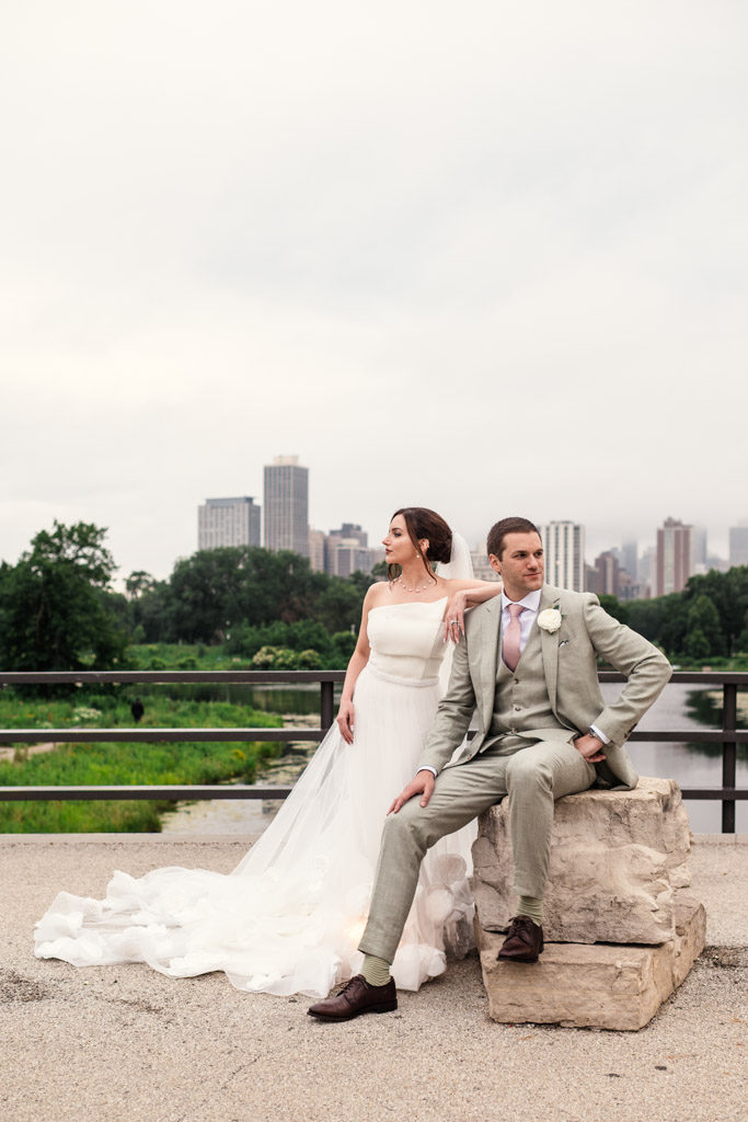 Bride in white gown and groom in light suit pose outdoors on a Lincoln Park bridge after their Lily Pool elopement, Chicago skyline and greenery in background