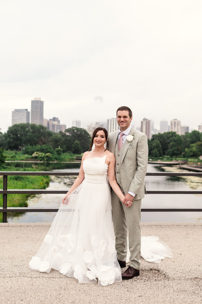 Portratit of bride in a white gown and groom in a light suit stand smiling by a Lincoln Park pond after their intimate Lily Pool elopement, Chicago skyline in the background