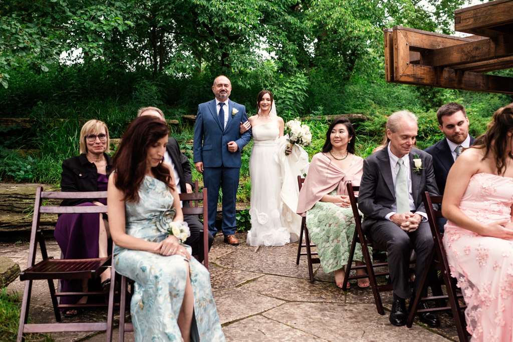 Bride in white dress and her father in blue suit walk down the aisle between seated guests at Alfred Caldwell Lily Pool elopement wedding ceremony