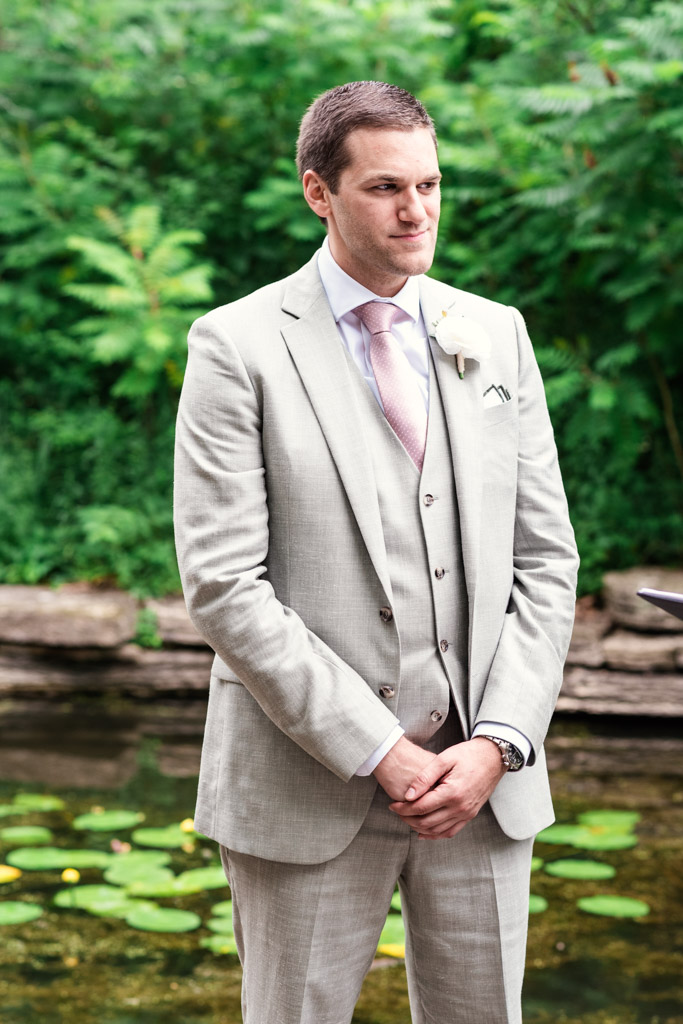 Groom in a light gray suit stands outdoors by Alfred Caldwell Lily Pool with lily pads, surrounded by lush green foliage during his elopement ceremony