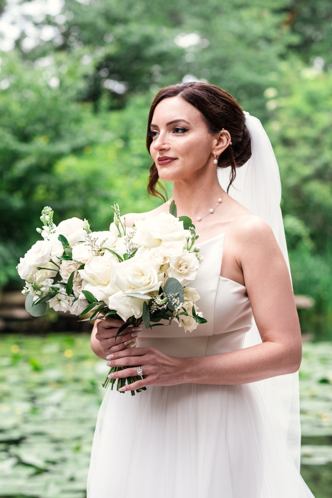 Bride in a white dress holds a bouquet of white flowers, standing outdoors by greenery and water during her Alfred Caldwell Lily Pool elopement
