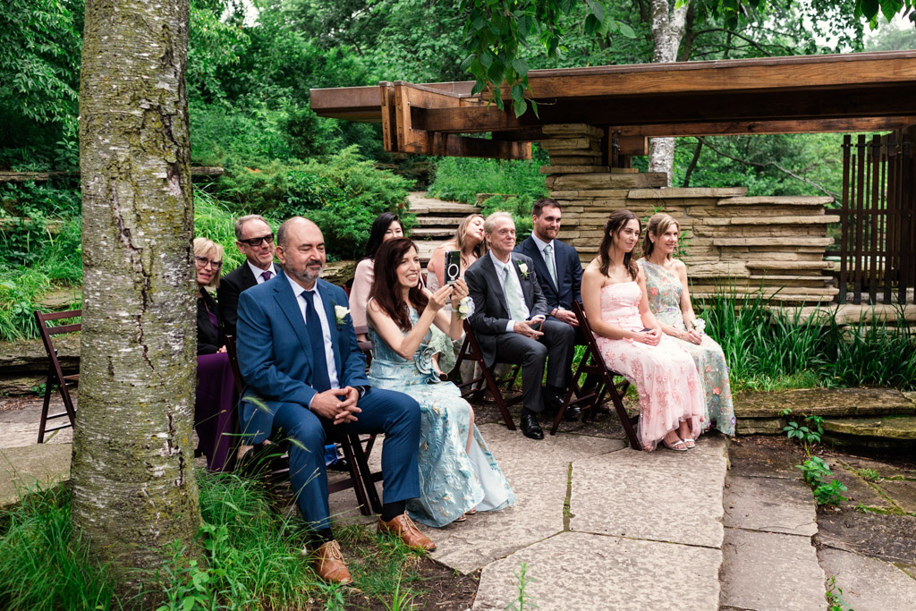 Guests in formal attire sit outdoors on folding chairs at Alfred Caldwell Lily Pool elopement wedding ceremony surrounded by greenery