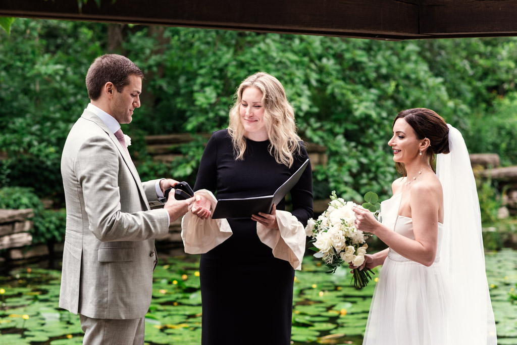 A bride and groom exchange rings during their Alfred Caldwell Lily Pool elopement, with an officiant between them