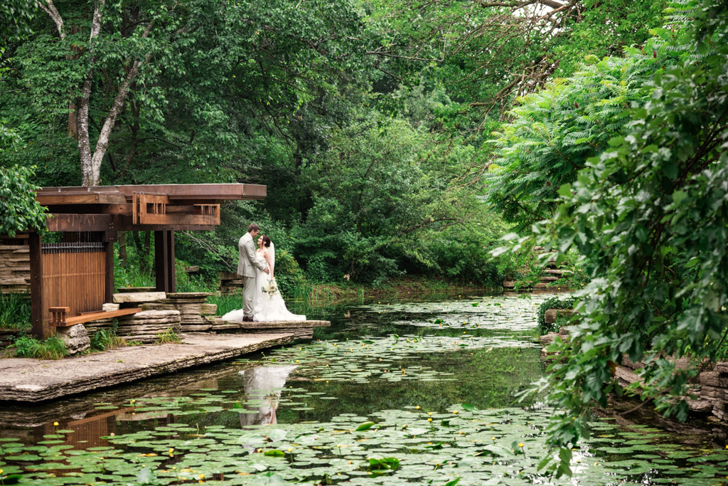A bride and groom stand together by Alfred Caldwell Lily Pool with lily pads, surrounded by lush green trees and plants