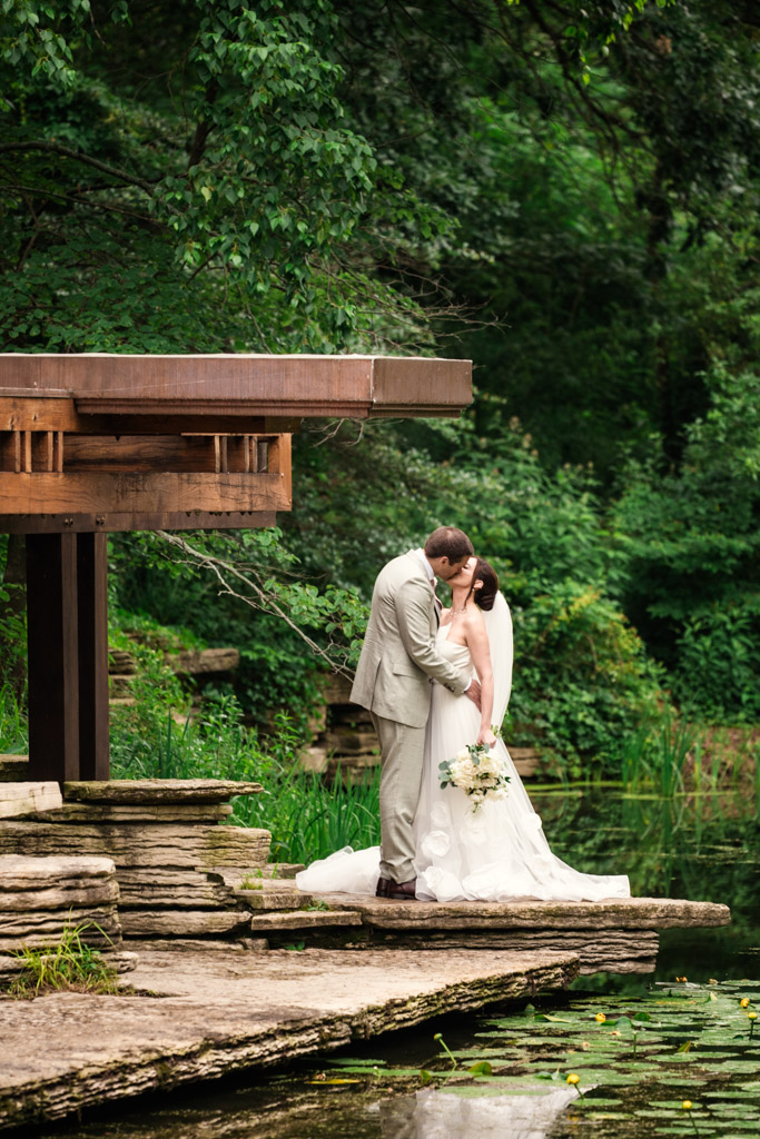 Bride and groom kiss by a modern wooden structure, surrounded by lush greenery during their Alfred Caldwell Lily Pool elopement