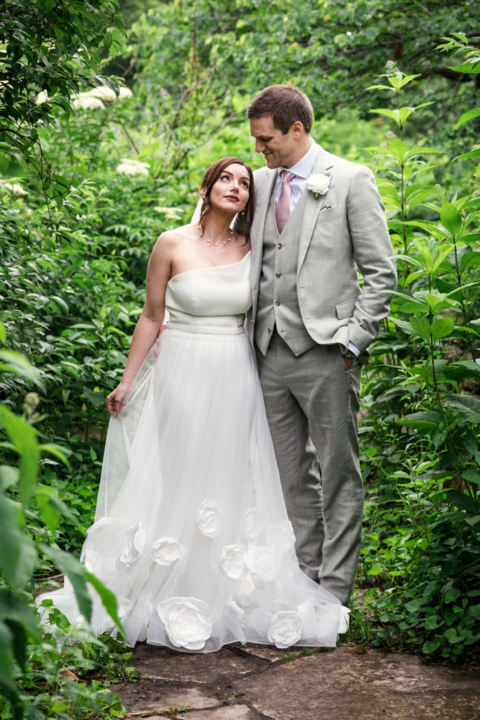 Bride and groom stand together on a Lincoln Park path, surrounded by lush green plants and trees, after their Alfred Caldwell Lily Pool elopement