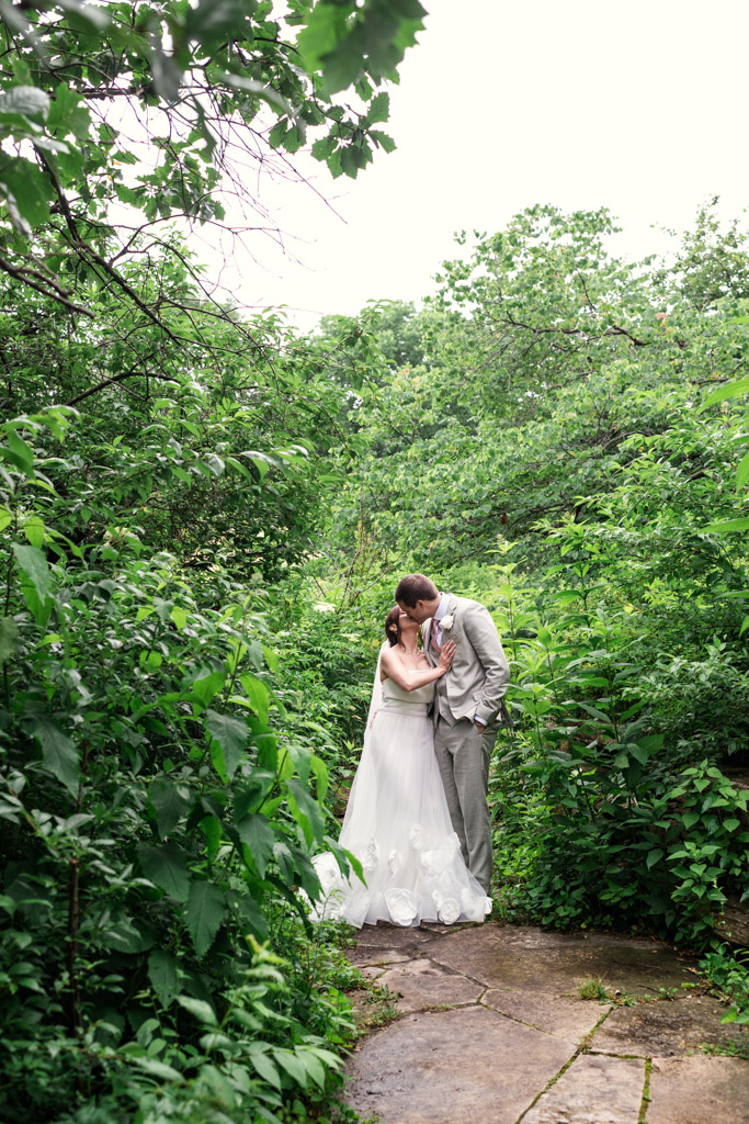 Bride and groom kiss on a stone Lincoln Park path surrounded by lush green foliage after their Lily Pool elopement