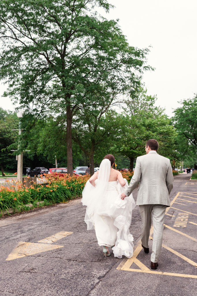 A bride and groom walk hand in hand through a tree-lined parking lot, seen from behind on a summer day during their Alfred Caldwell Lily Pool elopement