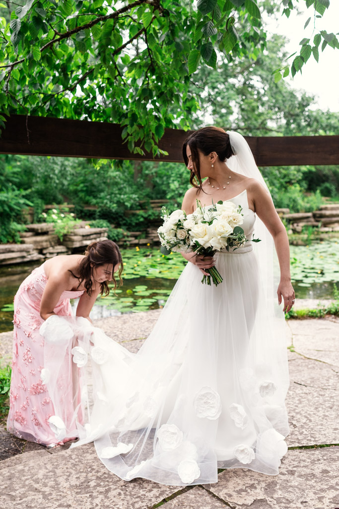Bride holding bouquet while bridesmaid adjusts her dress outdoors by Alfred Caldwell Lily Pool, surrounded by lush greenery