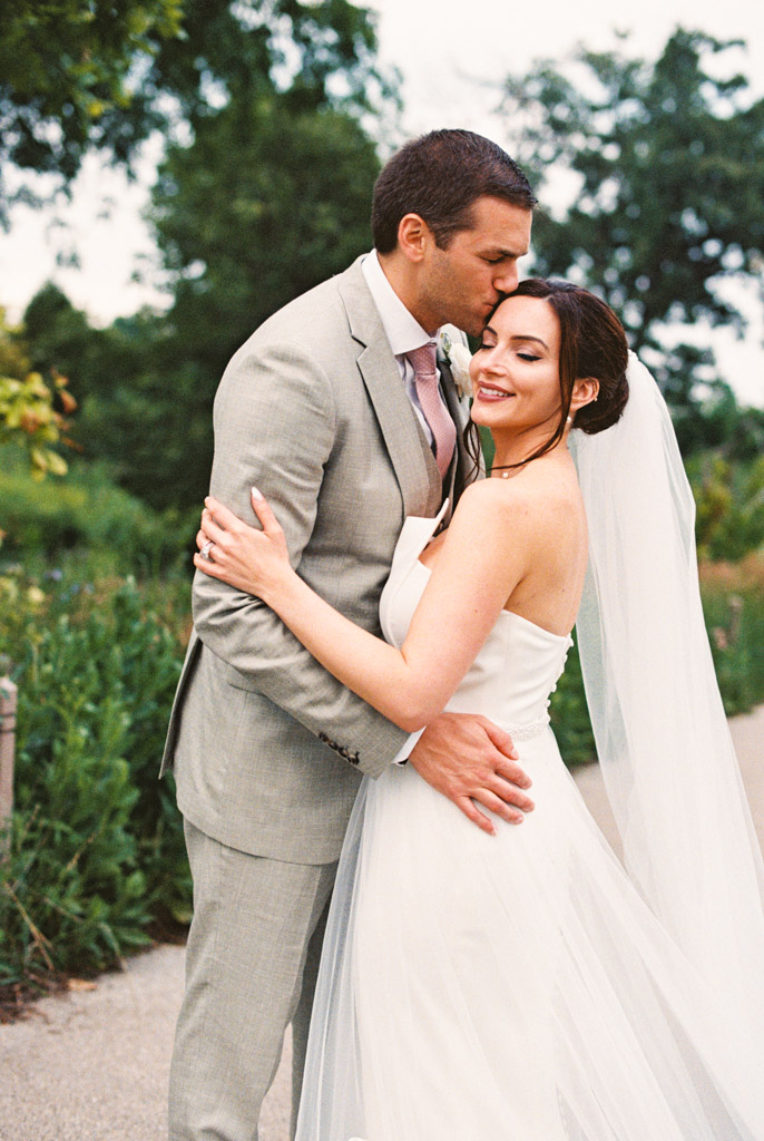 Film photo of groom in a light suit kissing happy bride in a white dress and veil in Lincoln Park after their intimate Lily Pool elopement