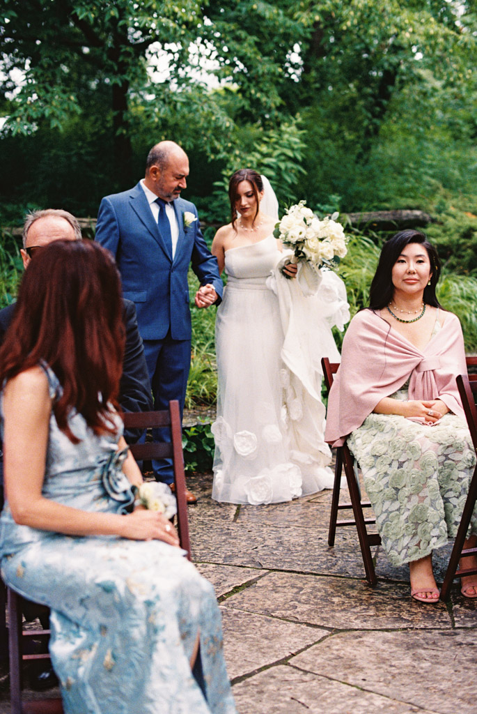 Film photo of bride in white dress walking down outdoor aisle with her father in a blue suit at her Alfred Caldwell Lily Pool elopement; guests sit and watch