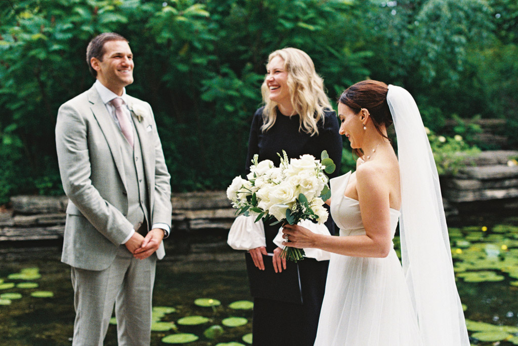 Film photo of bride in white dress holding bouquet, groom in suit, and officiant smiling outdoors by Alfred Caldwell Lily Pool during their intimate elopement ceremony