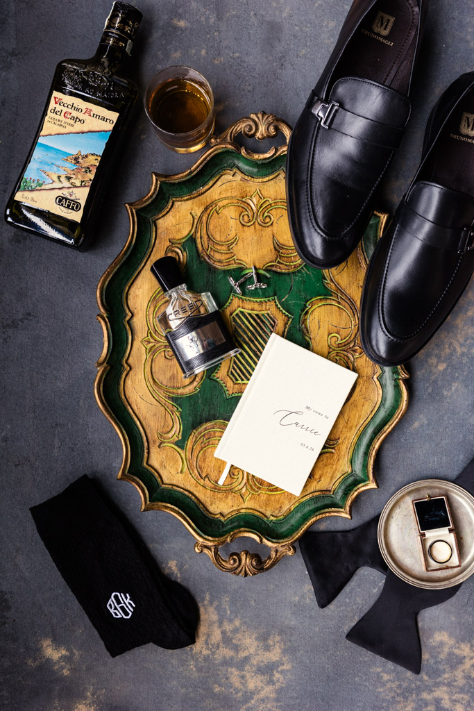 Flat lay of dress shoes, socks, cologne, liquor, bow tie, notebook, and glass on a decorative tray for an elegant Italian Villa wedding