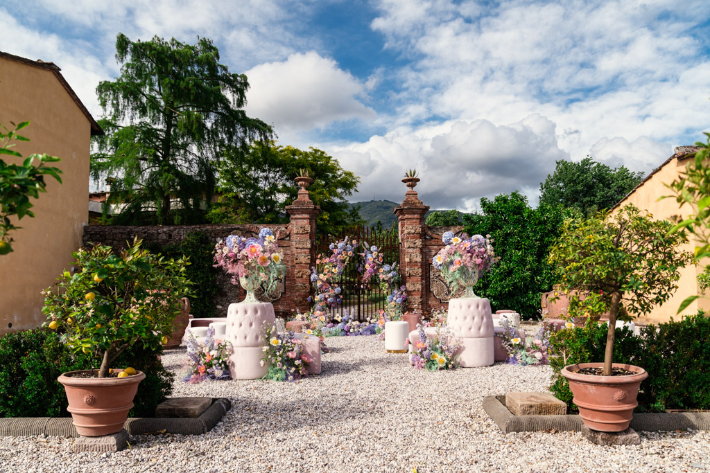Outdoor garden wedding ceremony space with pastel floral arrangements, white chairs, and potted plants under a partly cloudy sky outside an Italian Villa