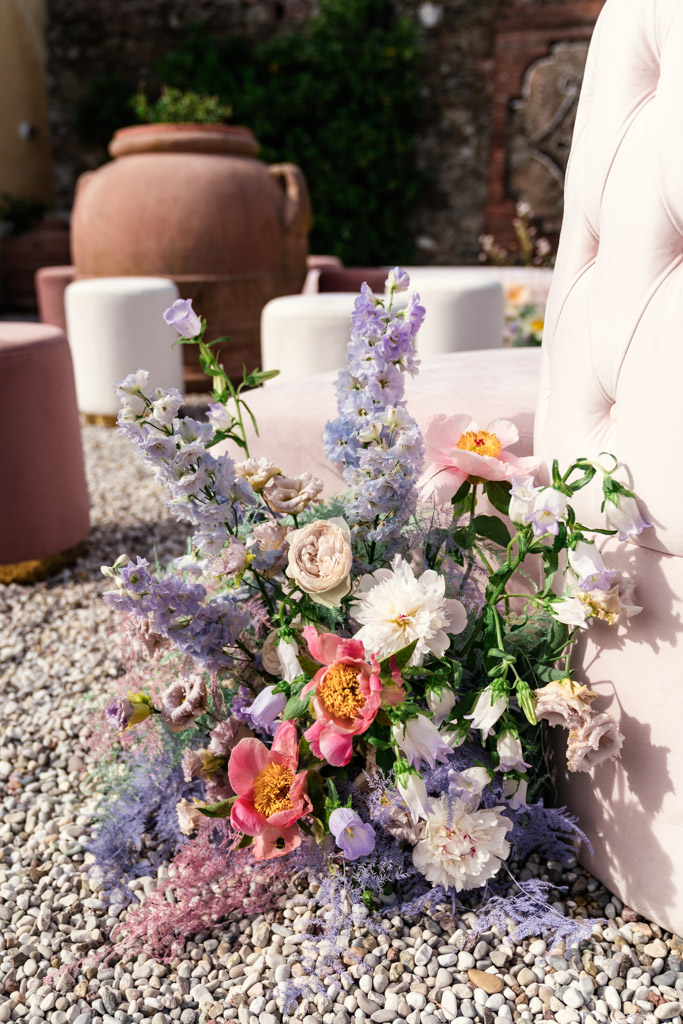 A bouquet of pink, white, and purple flowers rests on gravel near a pink, tufted chair and round ottomans for Italian Villa wedding celebration