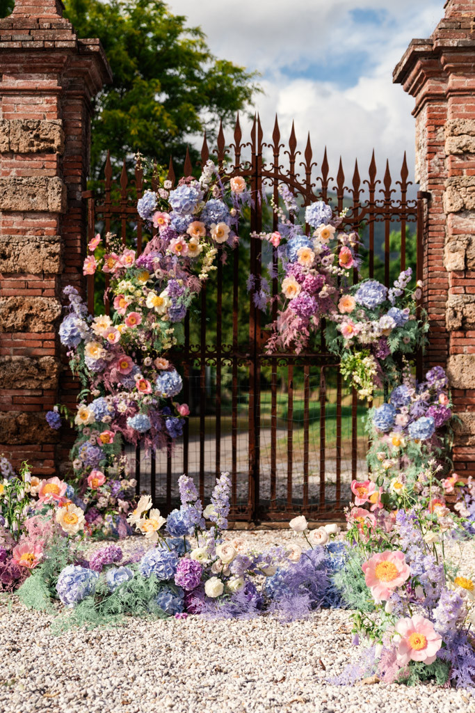 An ornate iron gate decorated with pastel flowers and greenery between brick pillars on a sunny day for Italian Villa wedding celebration