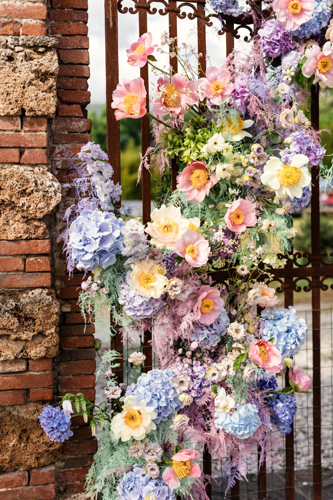Pastel flowers arranged on a rustic iron gate beside a brick wall, with greenery in the background for Italian Villa wedding celebration