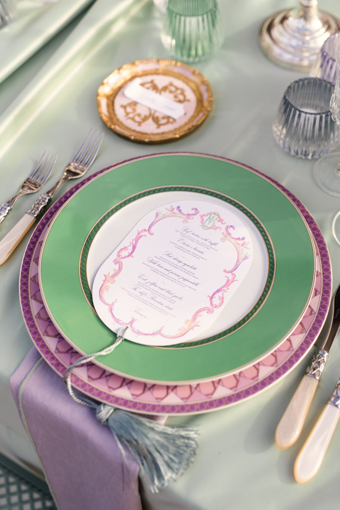 Guest table setting with layered green and purple plates, a menu, silverware, and glassware on a pastel tablecloth for an Italian Villa wedding celebration