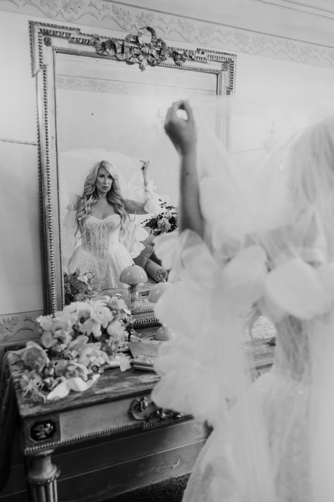 Black and white photo of bride adjusting her veil in front of a mirror, with flowers on a vintage table before her Italian Villa wedding ceremony