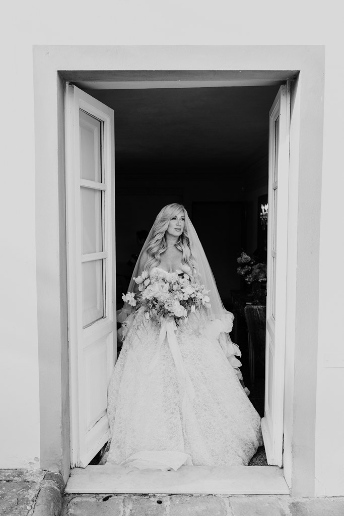 Black and white photo of bride in a lace gown and veil stands in a doorway of an Italian Villa wedding, holding a bouquet of flowers, looking to the side