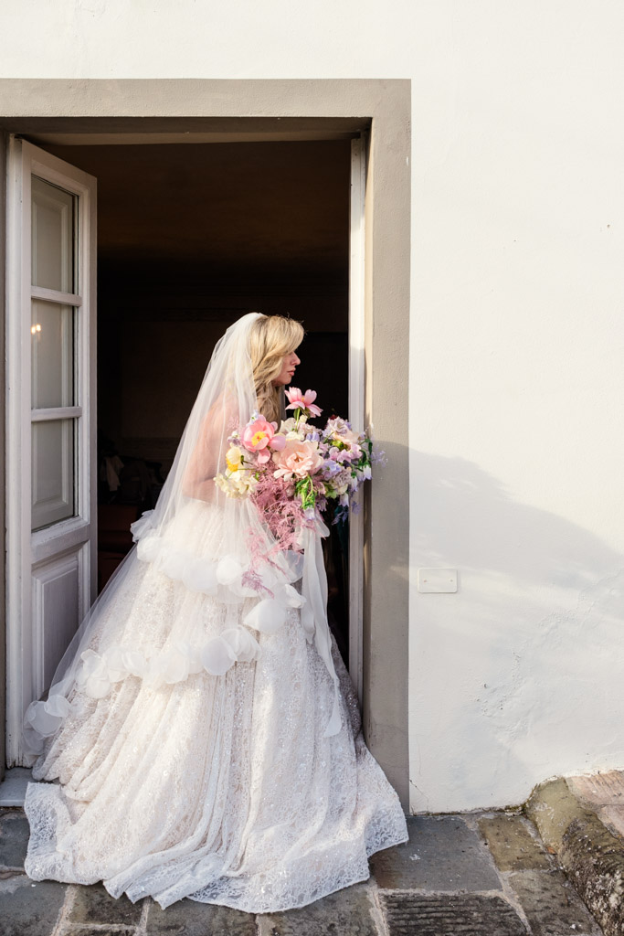 Bride in a white dress and veil holding a bouquet, standing in a sunlit doorway of an Italian Villa