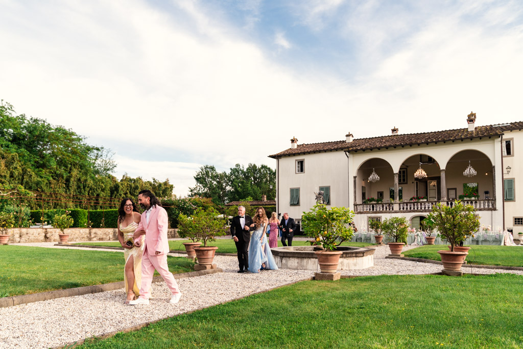 Wedding guests in formal attire walk on a gravel path in a garden by a large Italian Villa
