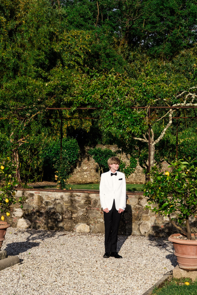 Bride's son in a white tuxedo stands on a pebble path in a sunny garden with green trees and stone walls before an Italian Villa wedding ceremony