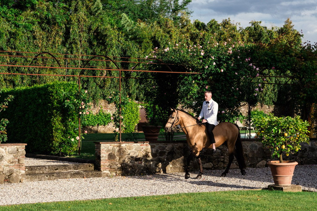 Groom rides a brown horse along a gravel path in a lush, green garden with stone walls during his Italian Villa wedding ceremony