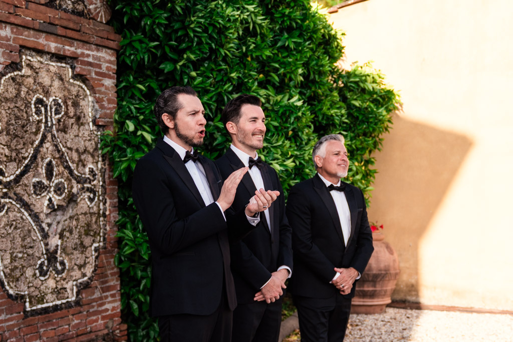 Three groomsmen in tuxedos stand outdoors by a brick wall and greenery at an elegant Italian Villa wedding ceremony