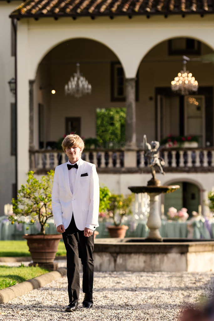 A young person in a white tuxedo walks on a gravel path in front of an elegant Italian Villa with arches and a fountain during Italian Villa wedding celebration in Tuscany