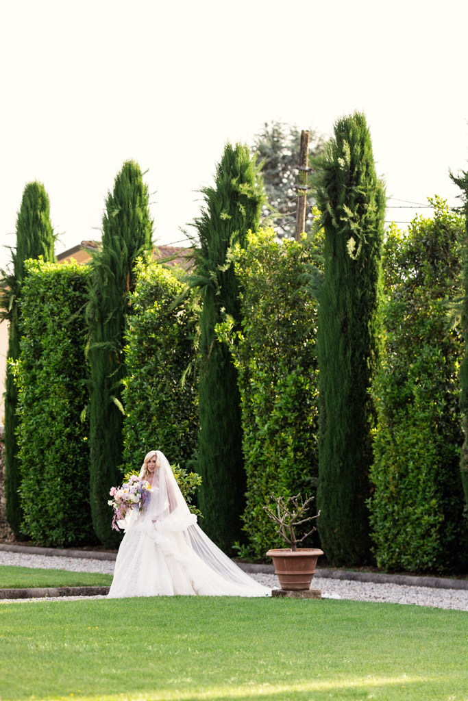 Bride in a white dress holds flowers, walking on a path near tall green hedges and trees, during her Italian Villa wedding