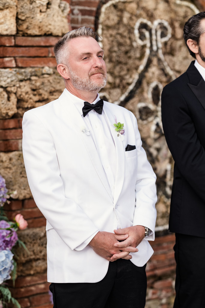 Groom in a white tuxedo jacket stands outdoors, hands clasped, with a stone wall and flowers behind him during his Italian Villa wedding ceremony