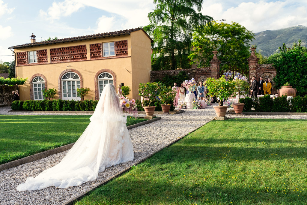 A bride in a white gown walks down a garden path toward wedding guests during her Italian Villa wedding ceremony