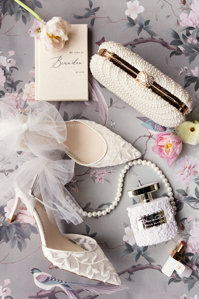 Bridal shoes, perfume, a pearl clutch, vow book, and flowers are beautifully arranged on floral paper for Italian Villa wedding