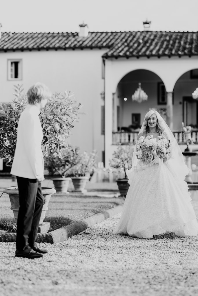 Black and white photo of bride walking toward a her son outdoors, both smiling, in front of an elegant Italian Villa wedding in Tuscany