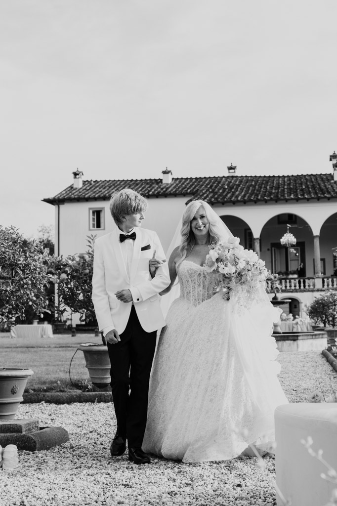 Black and white photo of bride in a white dress outside arm-in-arm with her son in a white tuxedo jacket at her Italian Villa wedding ceremony